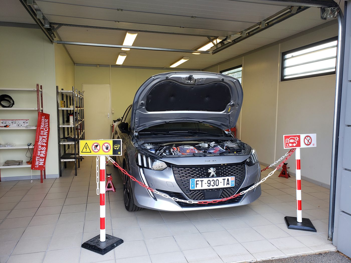 Un véhicule électrique capot ouvert dans un atelier, entouré d’une chaîne de balisage rouge et blanche et de panneaux « Limite de la zone de travail, ne pas franchir » et de risque électrique.